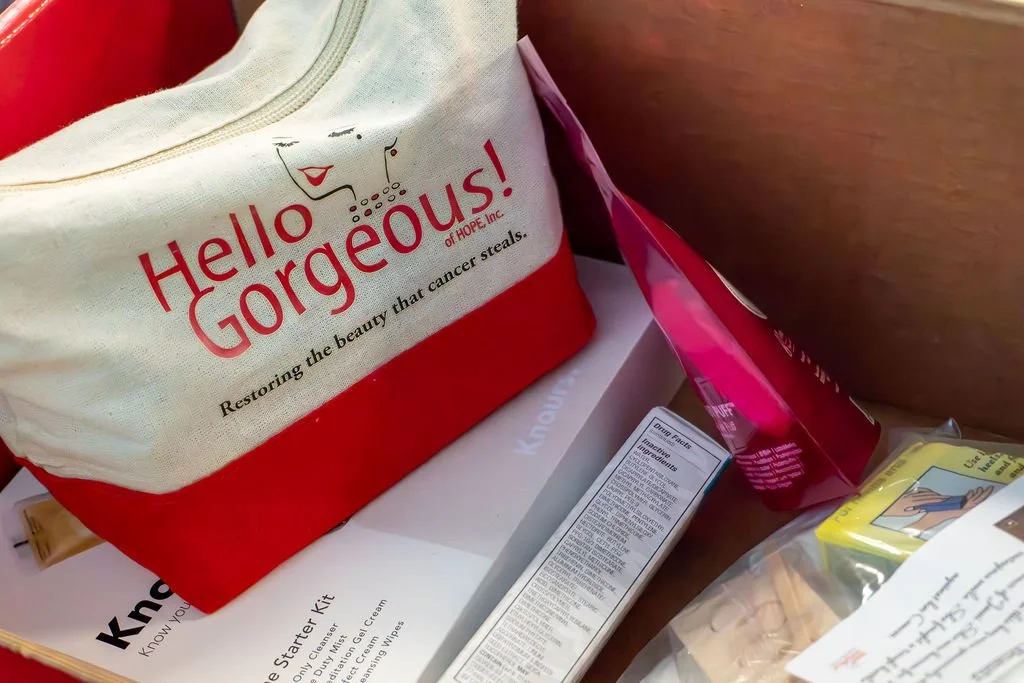 A tote bag with the words "Hello Gorgeous!" and a quote about cancer appears on a counter, surrounded by boxes and packages.