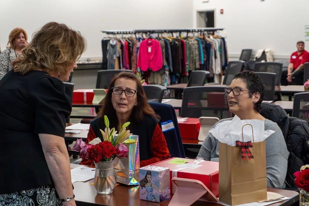 Women sitting at a table with gift bags and flowers, talking to a standing woman in a black top, in a room with hanging clothes in the background.
