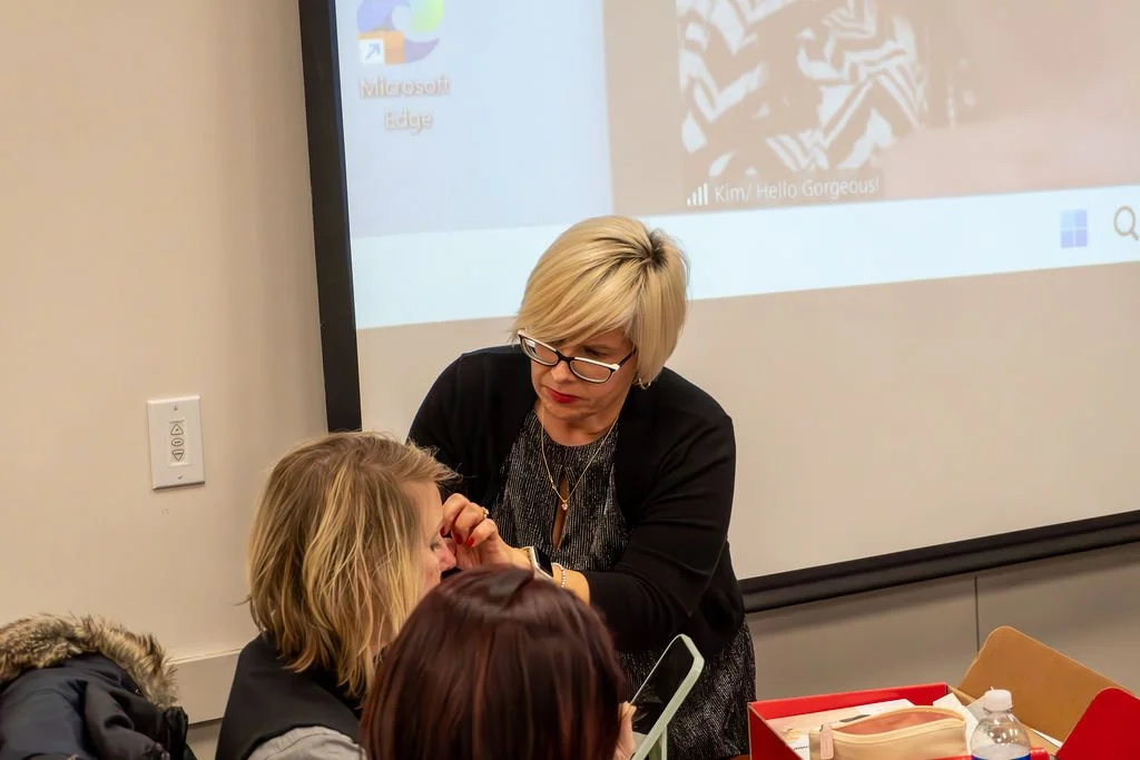 A woman with glasses and blonde hair applying makeup or face paint to a girl with blonde hair sitting at a table in a classroom or conference room. Other people are also seated nearby. A projector screen is visible in the background.