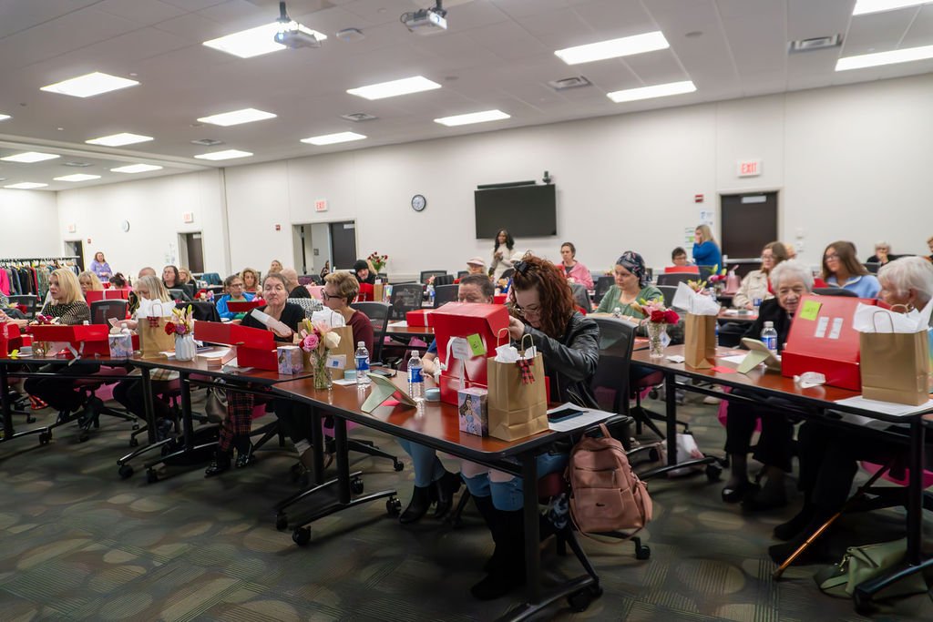 Women gathered in a conference room with gift boxes, bags, and flowers on tables, participating in a social or gift exchange event.