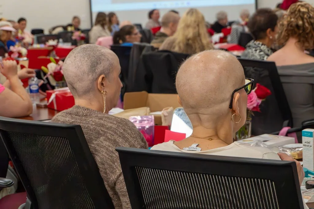 Women with shaved heads attending a conference or event, seated at tables decorated with flowers and gift boxes.