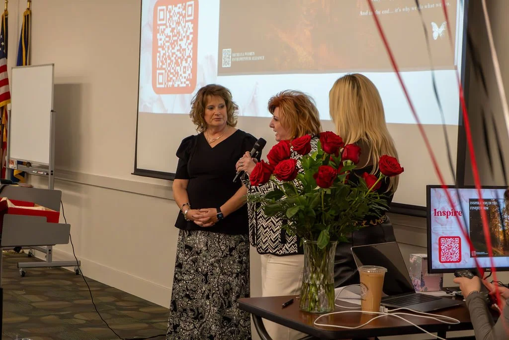 Three women standing and talking in a conference room, with a large vase of red roses on a desk in front of them and a projector screen in the background.