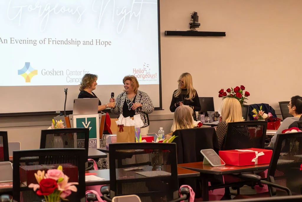 A woman on stage holding a microphone, smiling and talking to another woman in front of a projected screen at an event. Two other women and audience members are seated, with gift boxes and flowers on the tables. The screen has text about a women’s ev