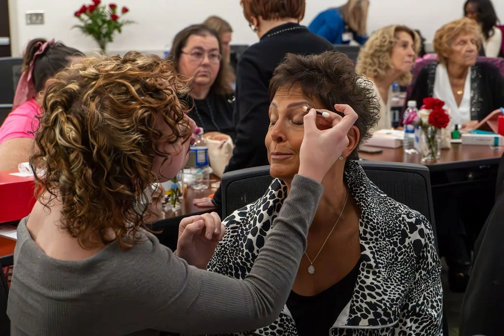 A woman is applying makeup to another woman in a busy room with other women seated at tables in the background.