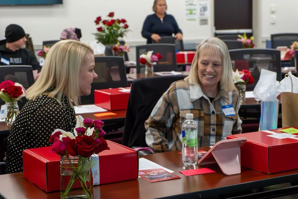 Two women sitting at a conference table, smiling and talking, with gifts, flowers, and a water bottle on the table, in a decorated meeting room.