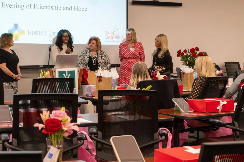 Women standing in front of a presentation screen during an event titled 'Evening of Friendship and Hope' at Goshhen Center, with seated attendees, flowers, gift bags, and a podium.