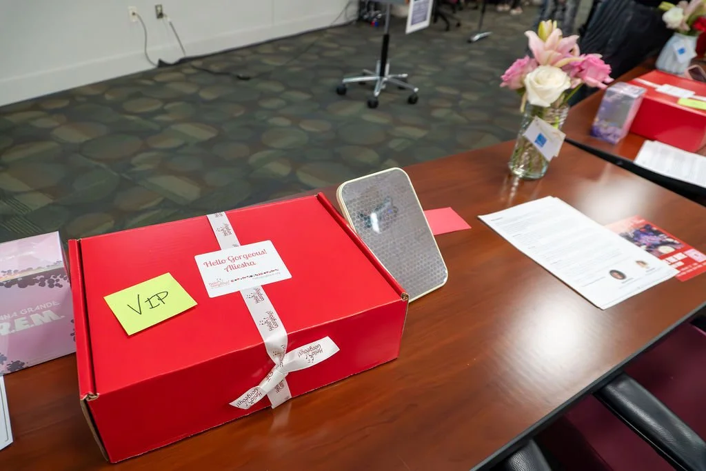 A red box labeled 'Hello Gorgeous Aisha' with a yellow sticky note 'VIP' on it, placed on a wooden table alongside papers, a small mirror, and a vase with pink and white flowers in an indoor setting.