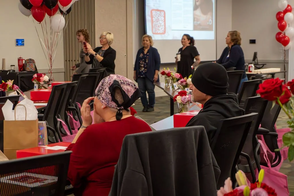 Women standing in front of a projected screen at a celebration with red and white balloons, flowers, and gift bags in the room.