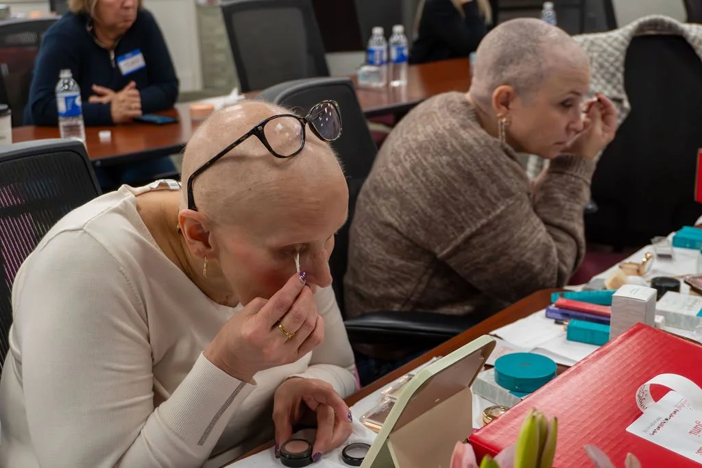A woman with a shaved head applying makeup at a table during a gathering or event, with makeup products and a mirror in front of her.