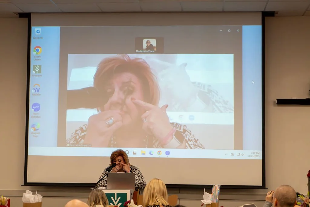 A woman is standing at a podium in front of a classroom with a projector screen behind her. The screen displays a large background image of a woman making a gagging face while pointing to her eye, with a dog in the background. The classroom has peopl