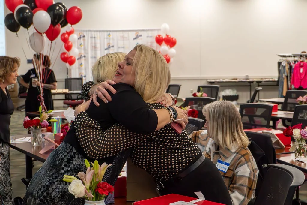 Women hugging at a celebration party with red, black, and white balloons, and decorated tables with flowers.
