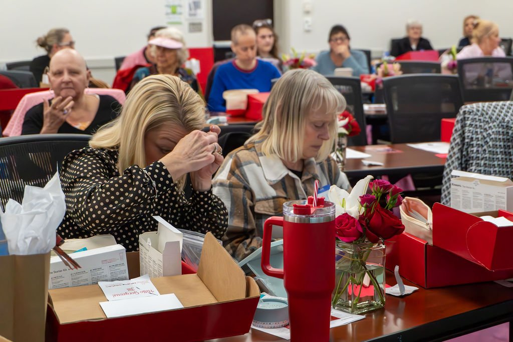A group of people seated at tables in a meeting room, with some holding their heads or looking down, surrounded by red gift boxes, flower arrangements, and various items.