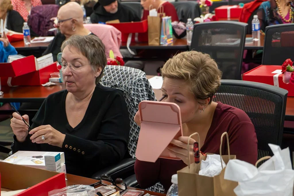 Women sitting at a decorated table, one is looking at her phone, and the other is applying makeup using a mirror. The table is decorated with red trays, gift bags, and gift boxes, indicating a celebration or gathering.