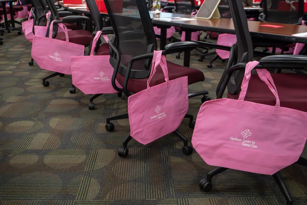 Pink tote bags hanging on black office chairs in a conference room, with tables and laptops, all bearing the logo of Goshen Center for Cancer Care.