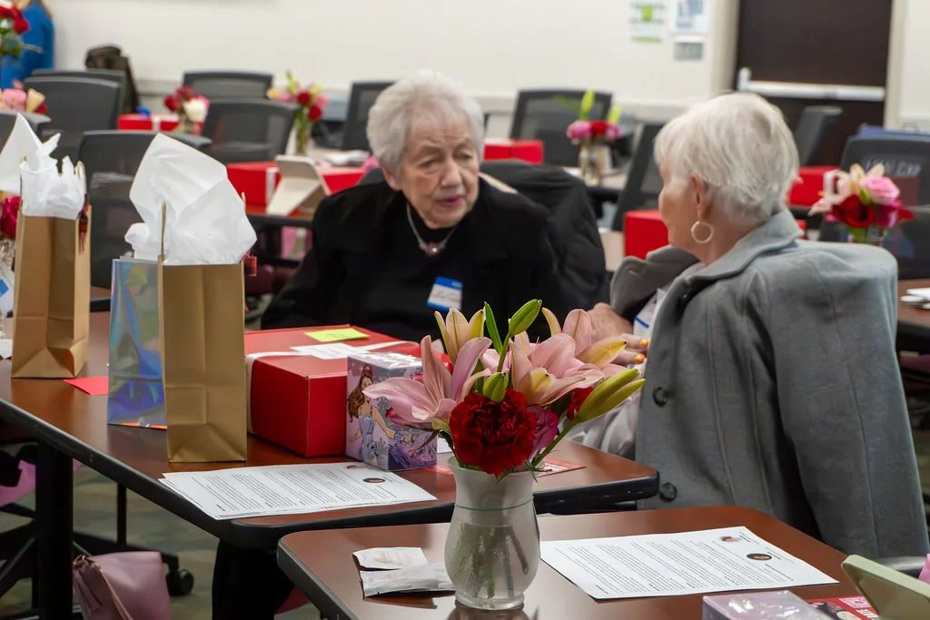 Two elderly women having a conversation at a conference table decorated with flowers and gift bags.