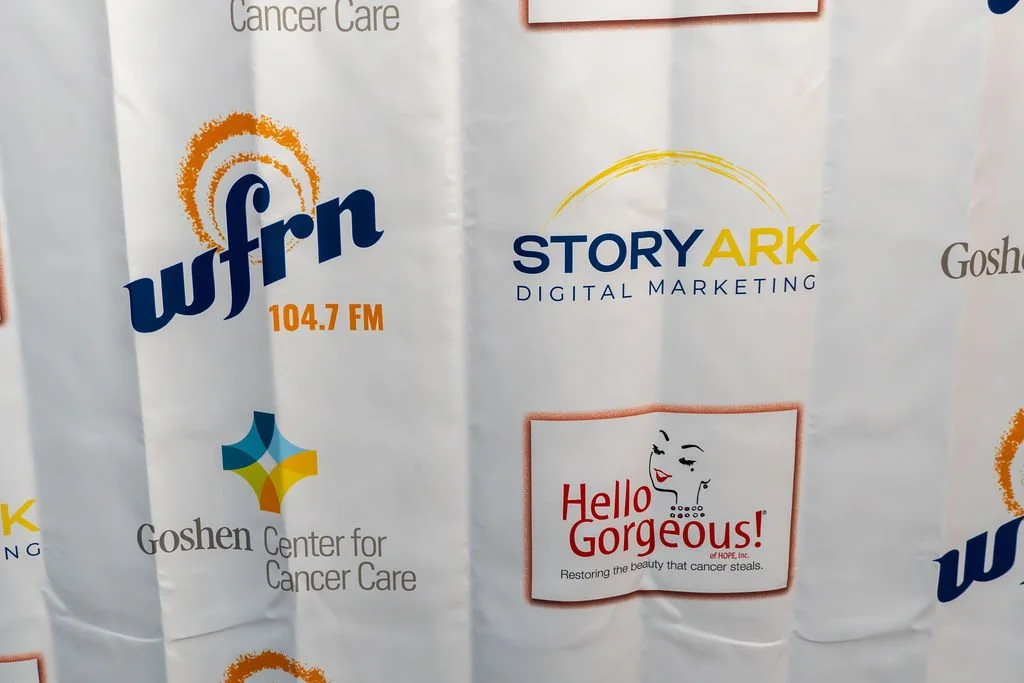 A promotional banner featuring logos of WFBN 104.7 FM, StoryArk Digital Marketing, Goshen Center for Cancer Care, and Hello Gorgeous! with a tagline about restoring beauty for cancer patients.