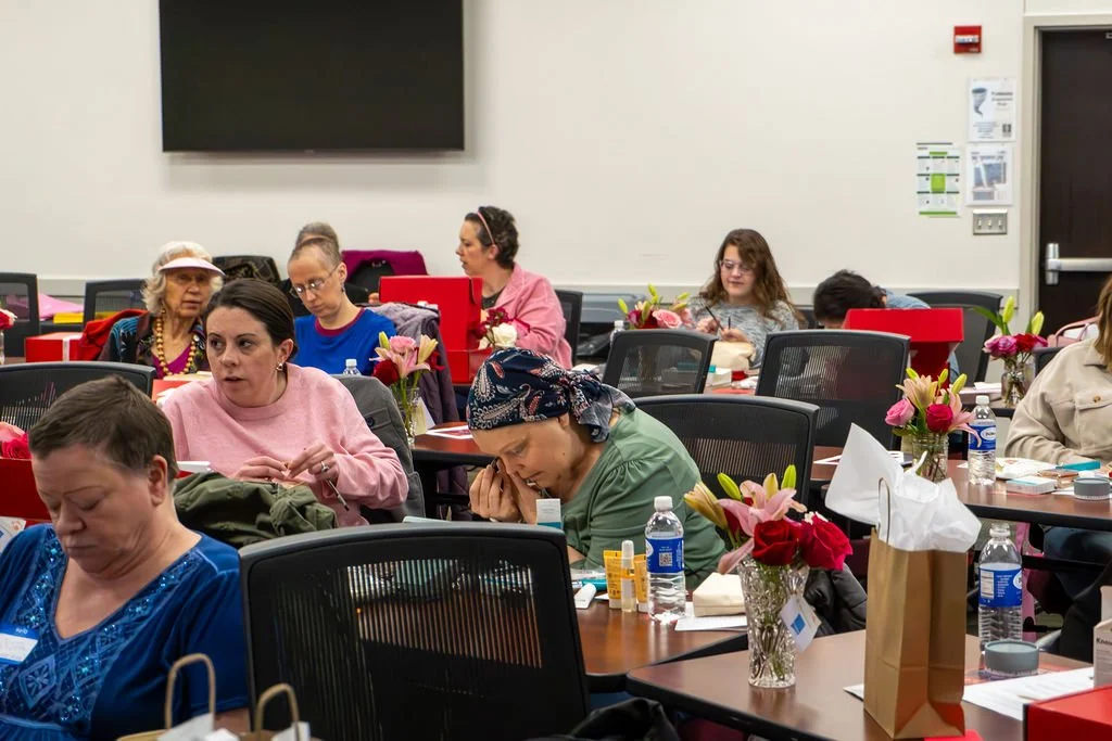 Several women are sitting at tables in a conference room with pink and red flower arrangements, water bottles, and conference materials, some appearing focused, listening, or taking notes.