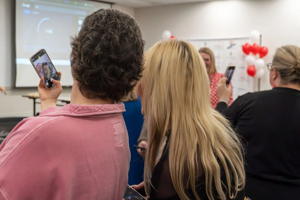 Group of people in a classroom taking photos with smartphones, with Valentine's Day balloons in the background.