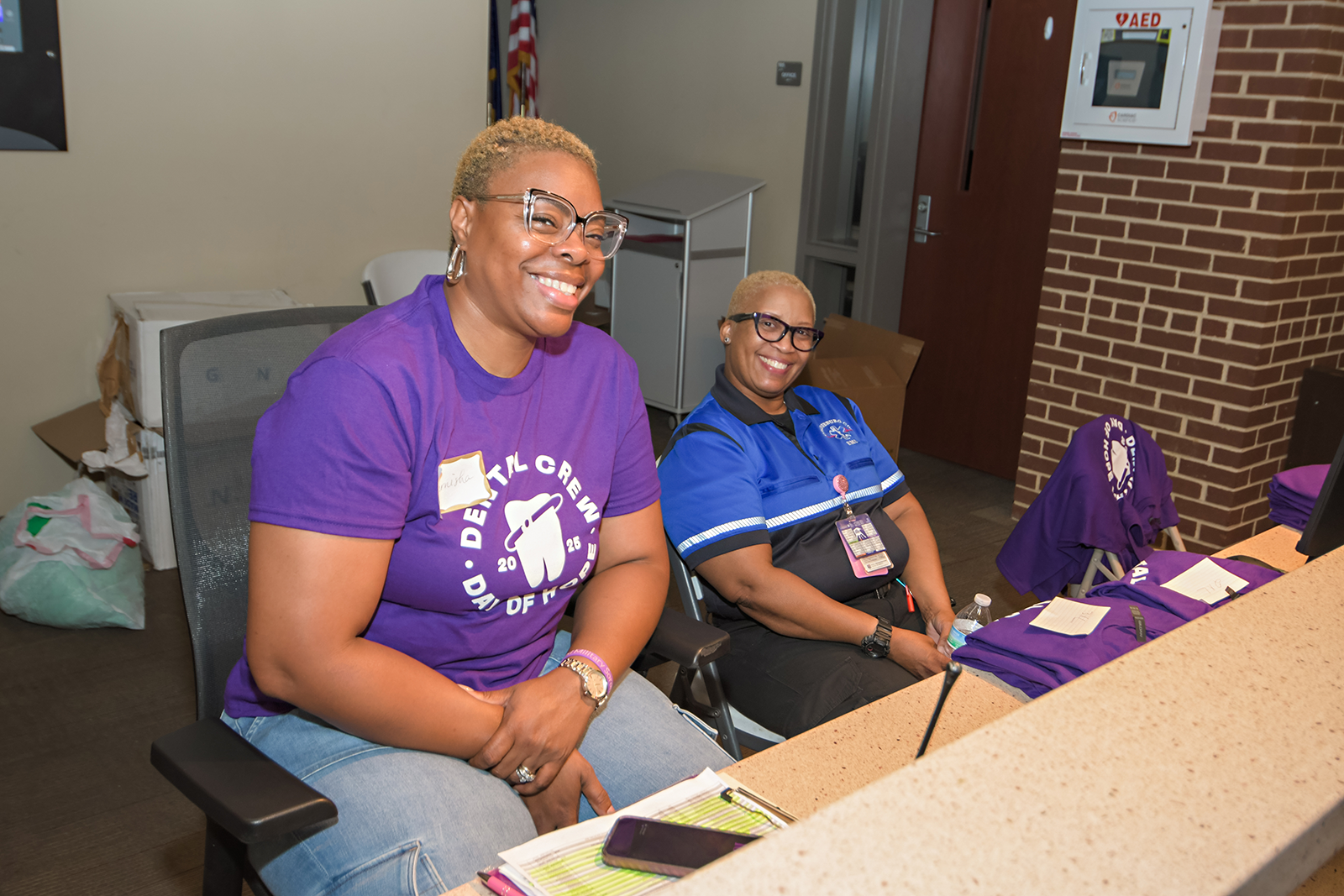 Two women sitting at a desk, smiling, with purple and blue shirts, in a room with a brick wall and a fire alarm panel.