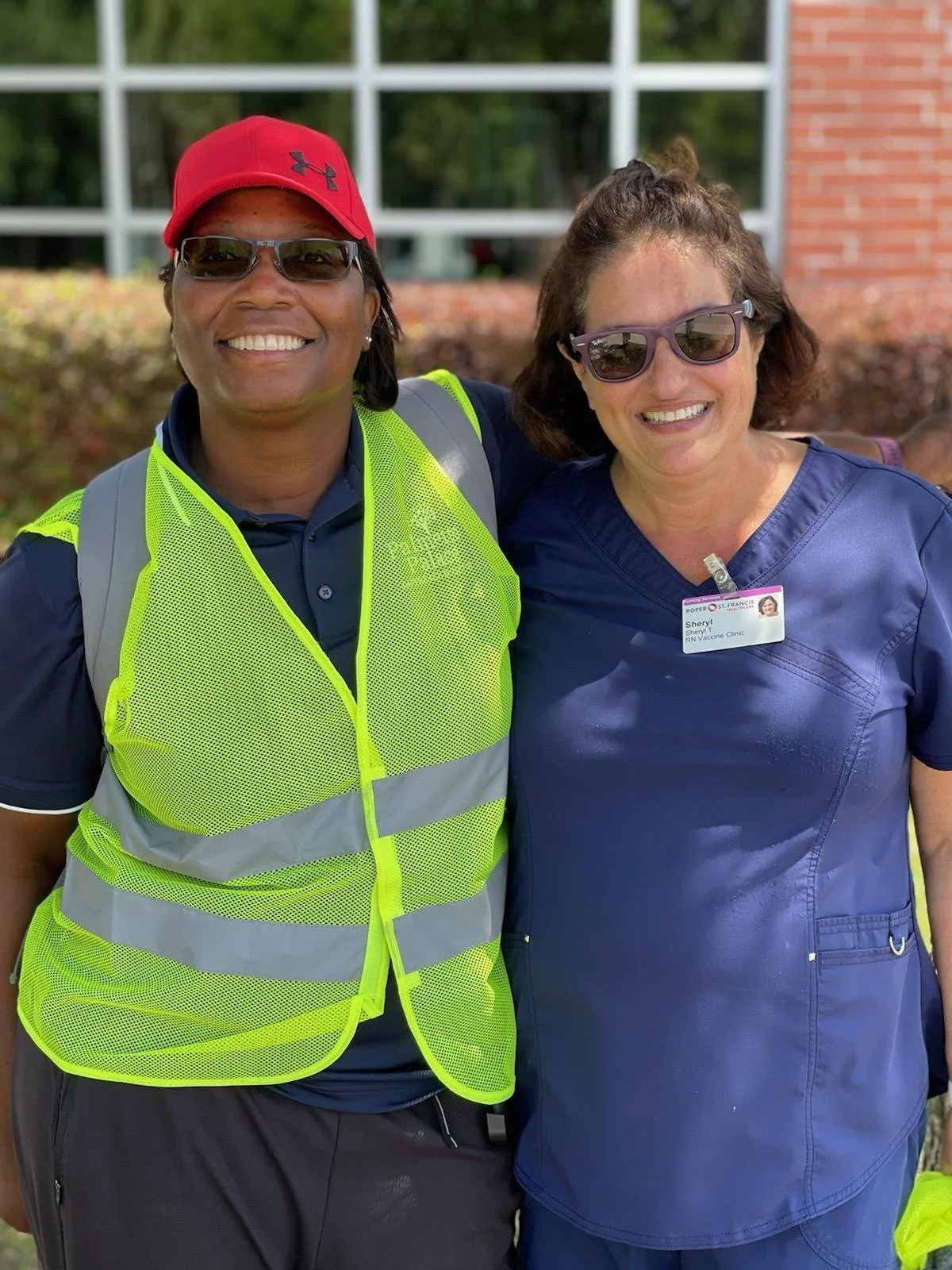 Two women standing outside smiling, one wearing a yellow safety vest and sunglasses, the other in blue scrubs with a nurse badge.