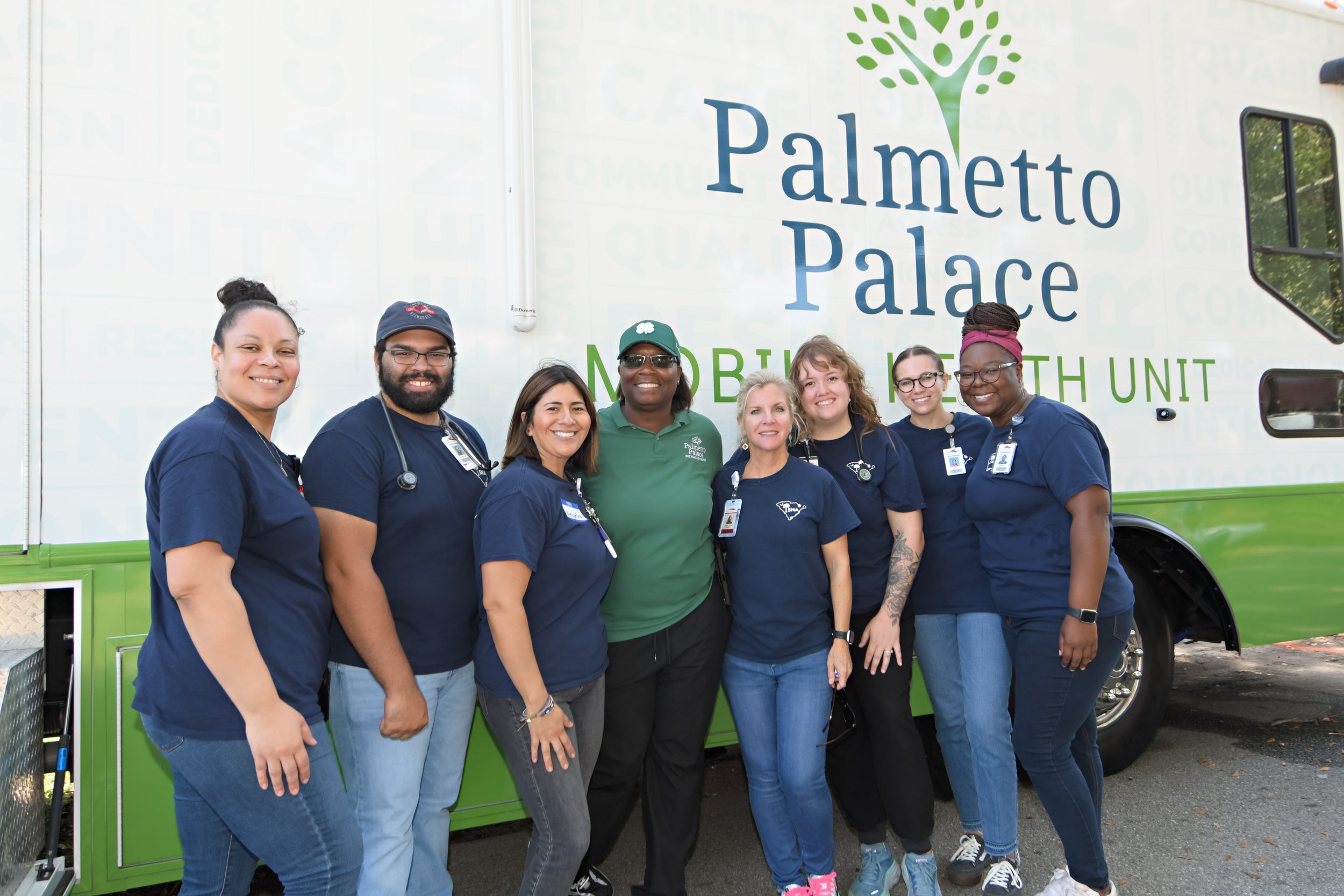 Group of healthcare professionals standing together in front of a mobile health unit with 'Palmetto Palace' written on it, outdoors during daytime.