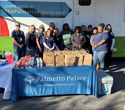 A mobile health unit in a grassy area with a group of people gathered near it, including a table with informational material and a large poster honoring Gilda Cobb-Hunter. The mobile health unit displays the Palmetto Palace logo and is shaped like a recreational vehicle with an awning extended.