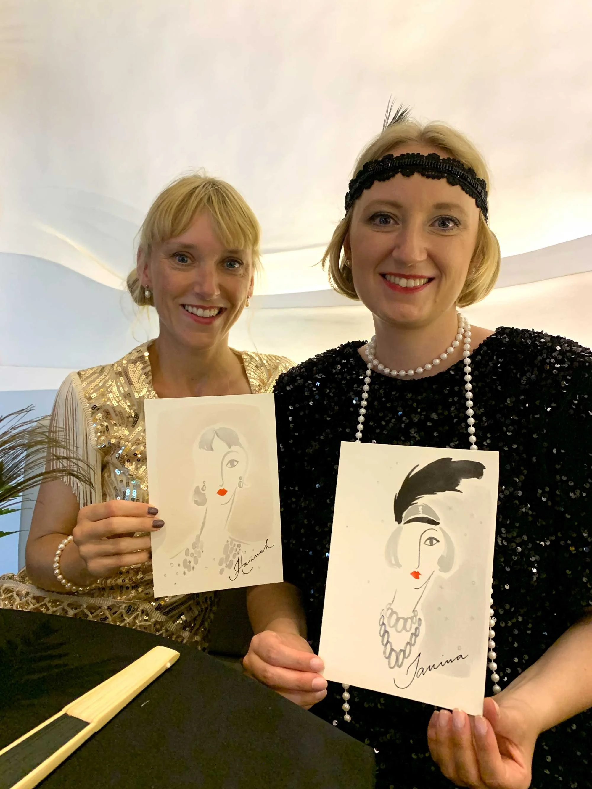 Two women in vintage-style dresses holding illustrated portraits of a woman with a hat and pearls, smiling at the camera.