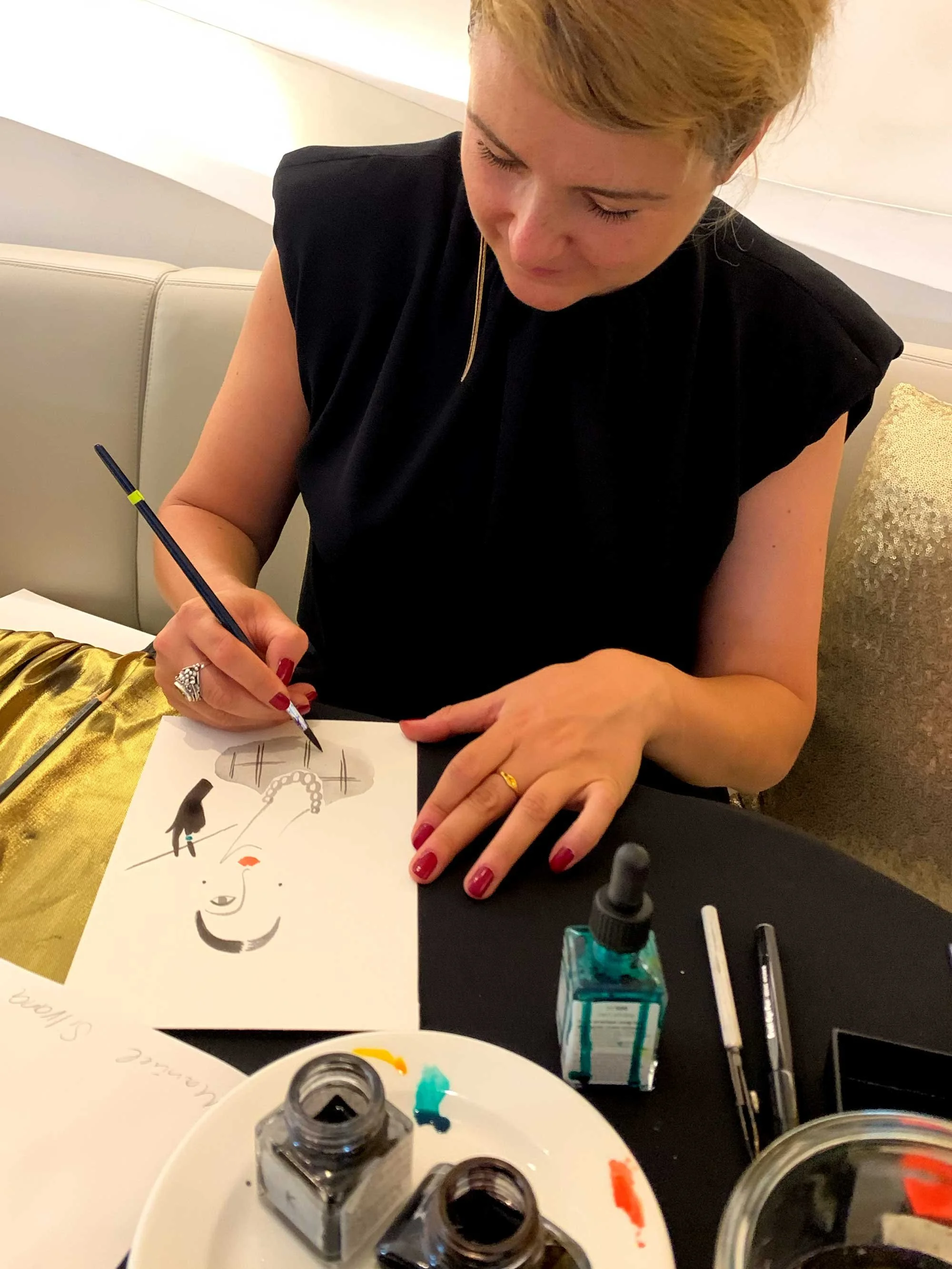 A woman with red hair and red nails drawing a portrait of a woman with makeup and jewelry on white paper at a table with ink bottles, pens, and paint, in a room with a beige couch and decorative pillows.