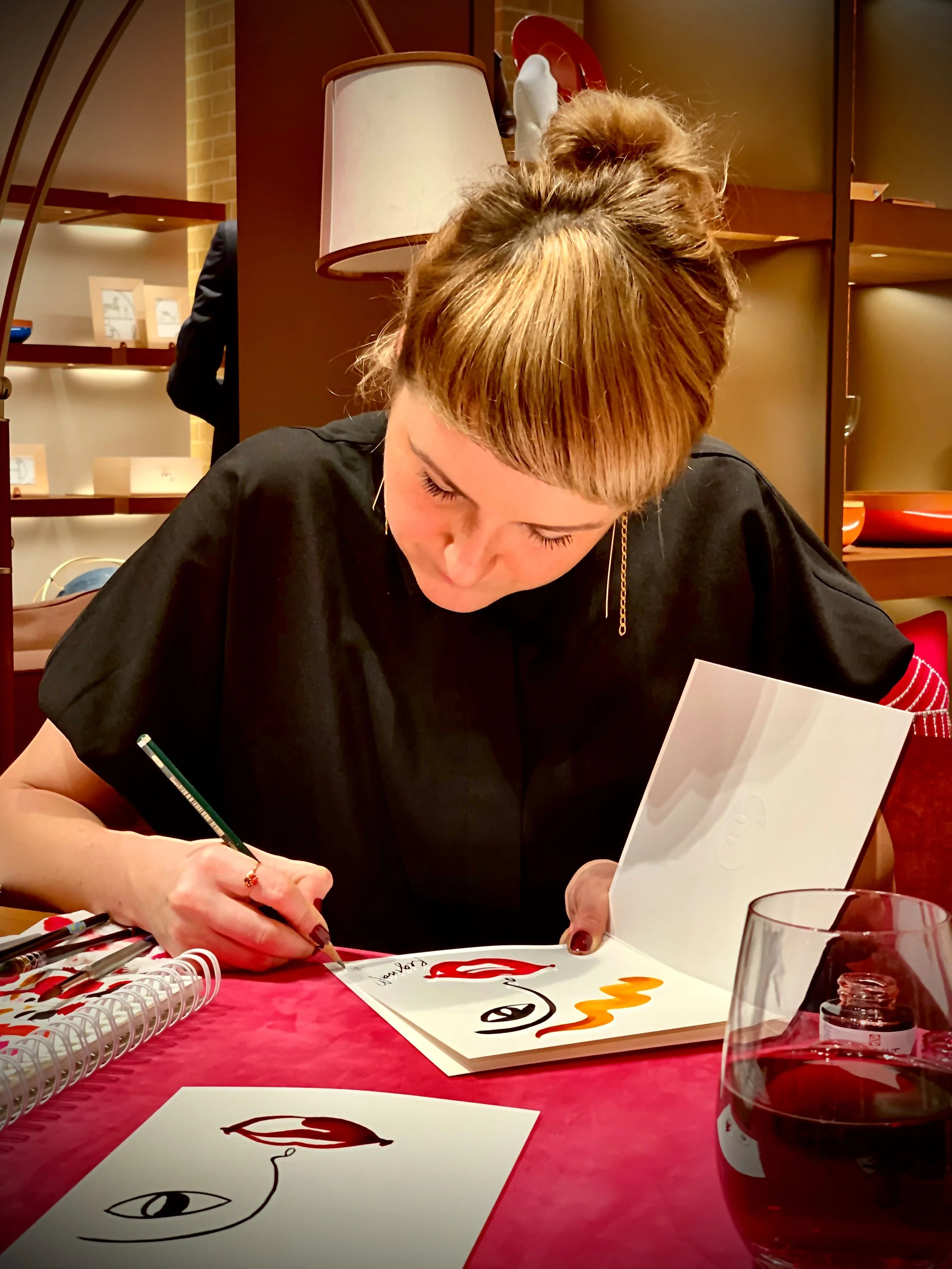 A woman with reddish hair styled in a bun, wearing a black shirt with red and white striped sleeves, is sitting at a pink table and signing a book with a green marker. There are artwork of lips and an eye drawn on the papers and book.