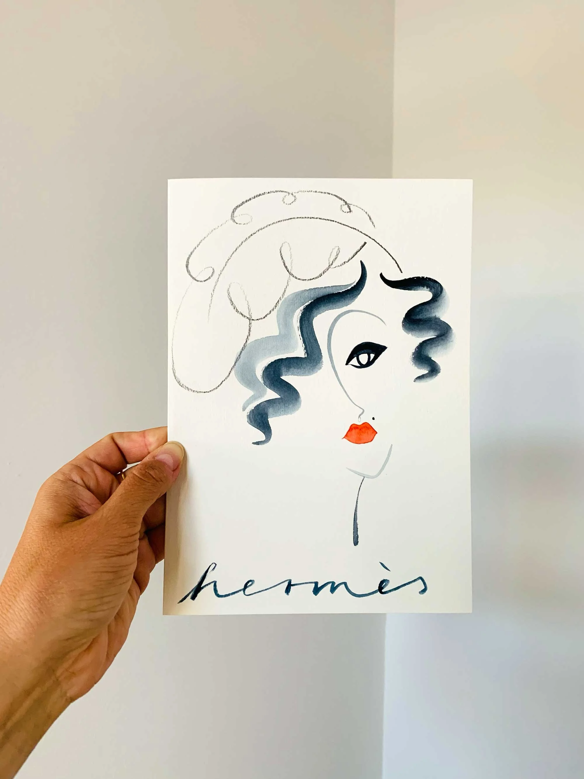 Hand holding an artistic sketch of a woman's face with curly hair, red lips, and a minimalistic style, with the word 'Hermès' written at the bottom.