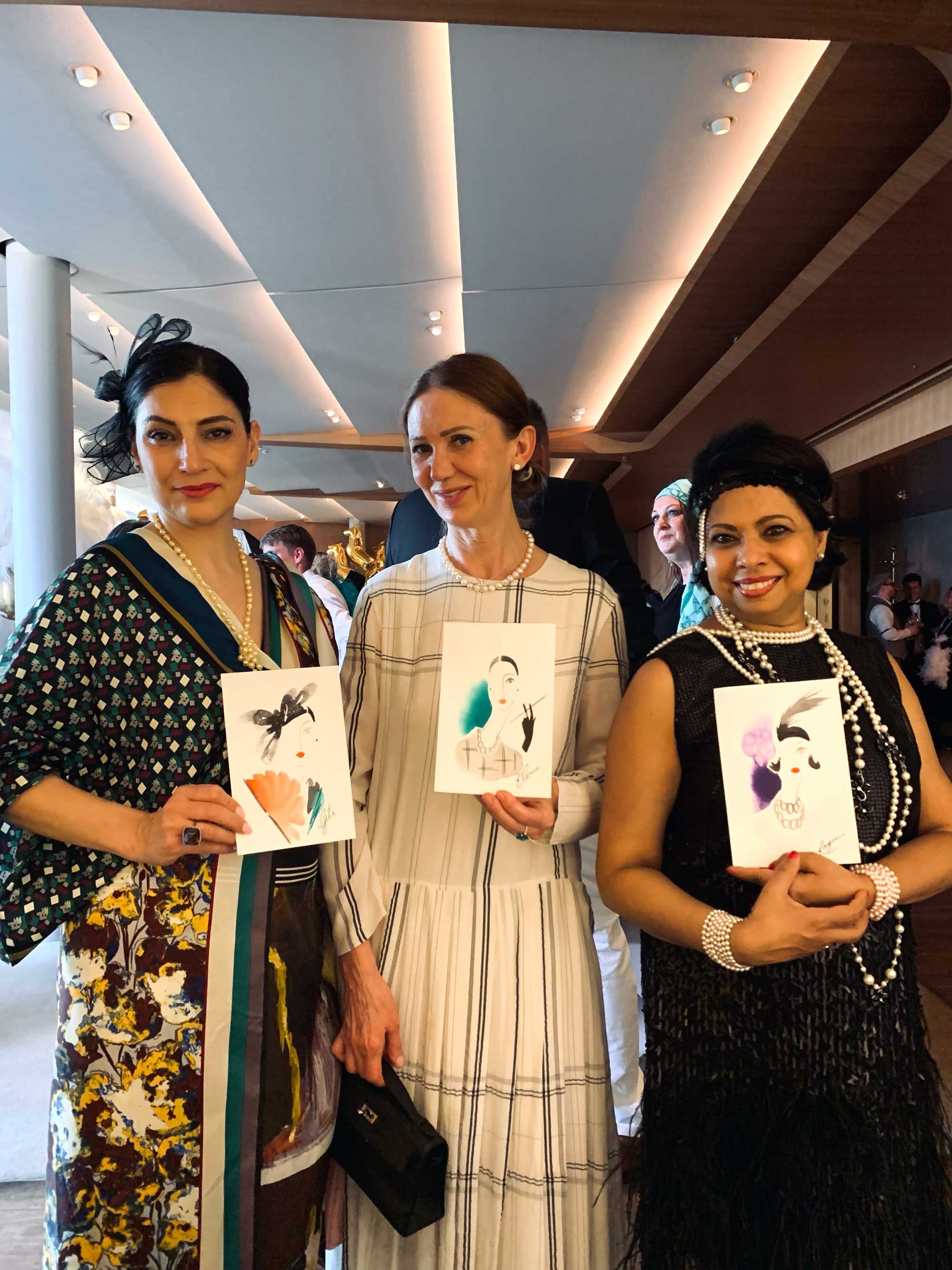 Three women dressed in vintage fashion from the 1920s or 1930s, holding illustrated portraits of women in vintage style, at an indoor event with a modern ceiling and warm lighting.