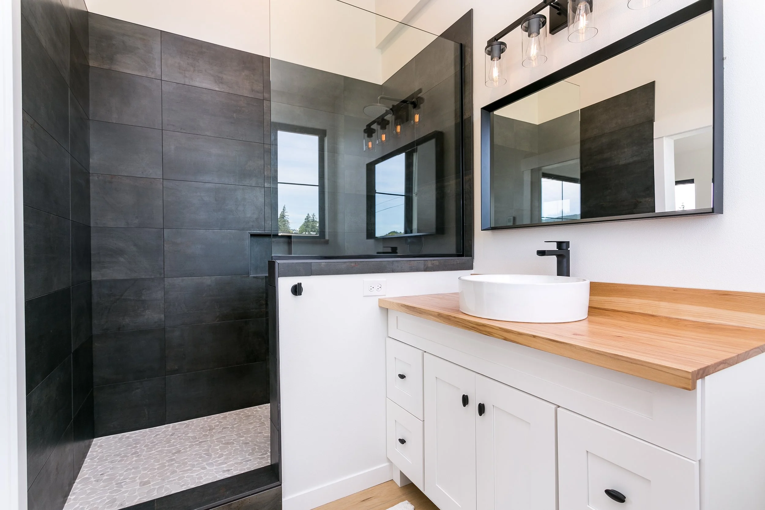 Modern bathroom with a white vanity, wooden countertop, black faucet, oval sink, large mirror, and a dark tiled walk-in shower.