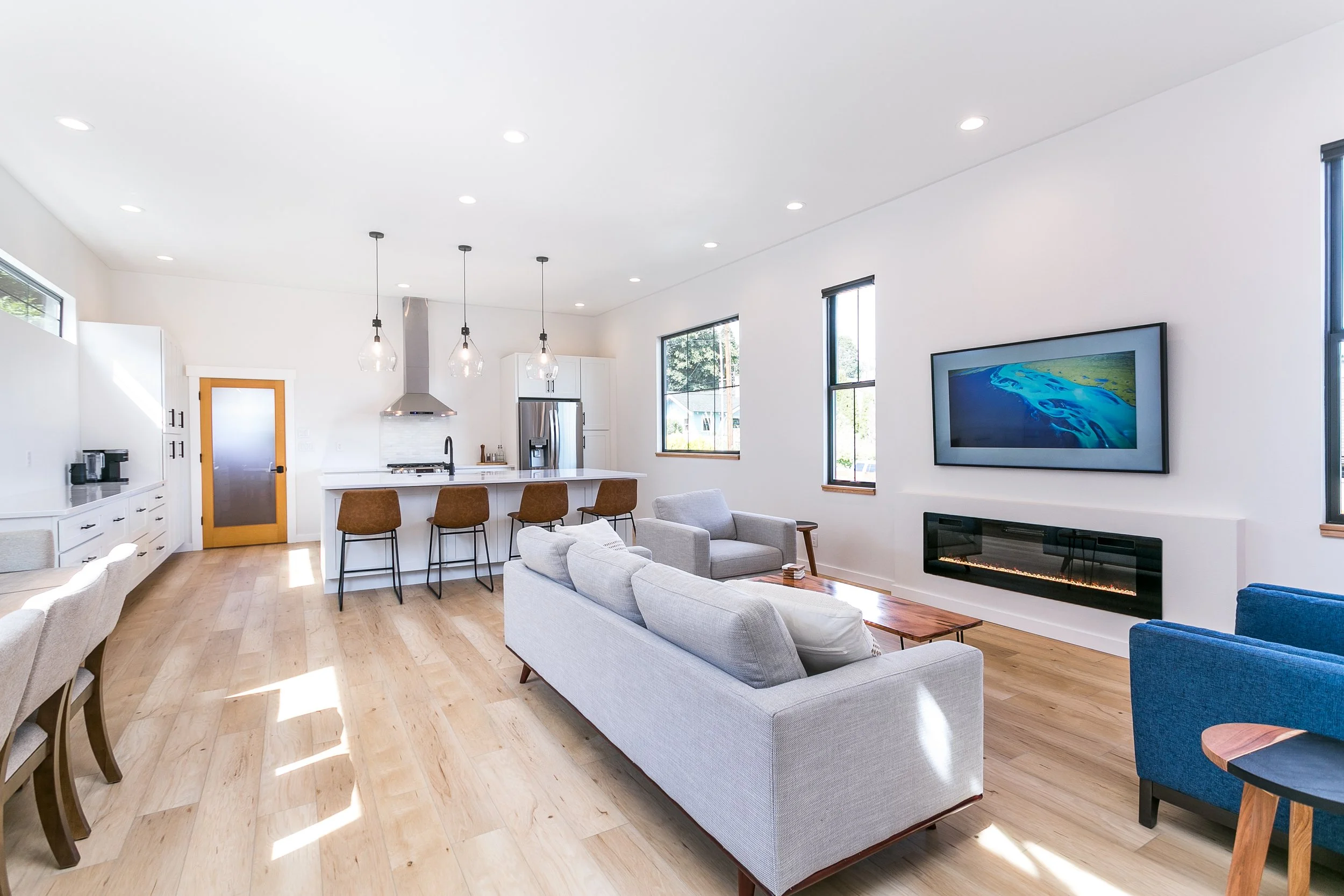 Modern open-concept living room and kitchen with light wood flooring, white walls, gray sofa, blue armchairs, wall-mounted TV, electric fireplace, kitchen island with bar stools, pendant lights, and stainless steel appliances.