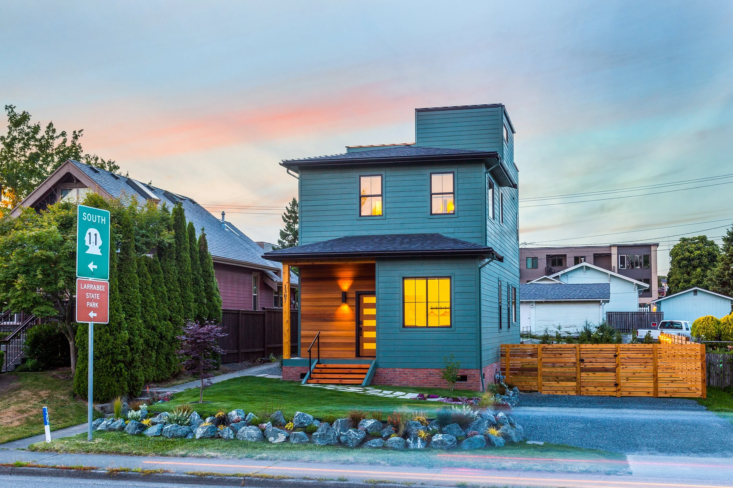 Modern blue house with illuminated windows at dusk, surrounded by a neat yard, wooden fence, and road signage directing to Larrabee State Park, in a residential neighborhood with other houses visible.