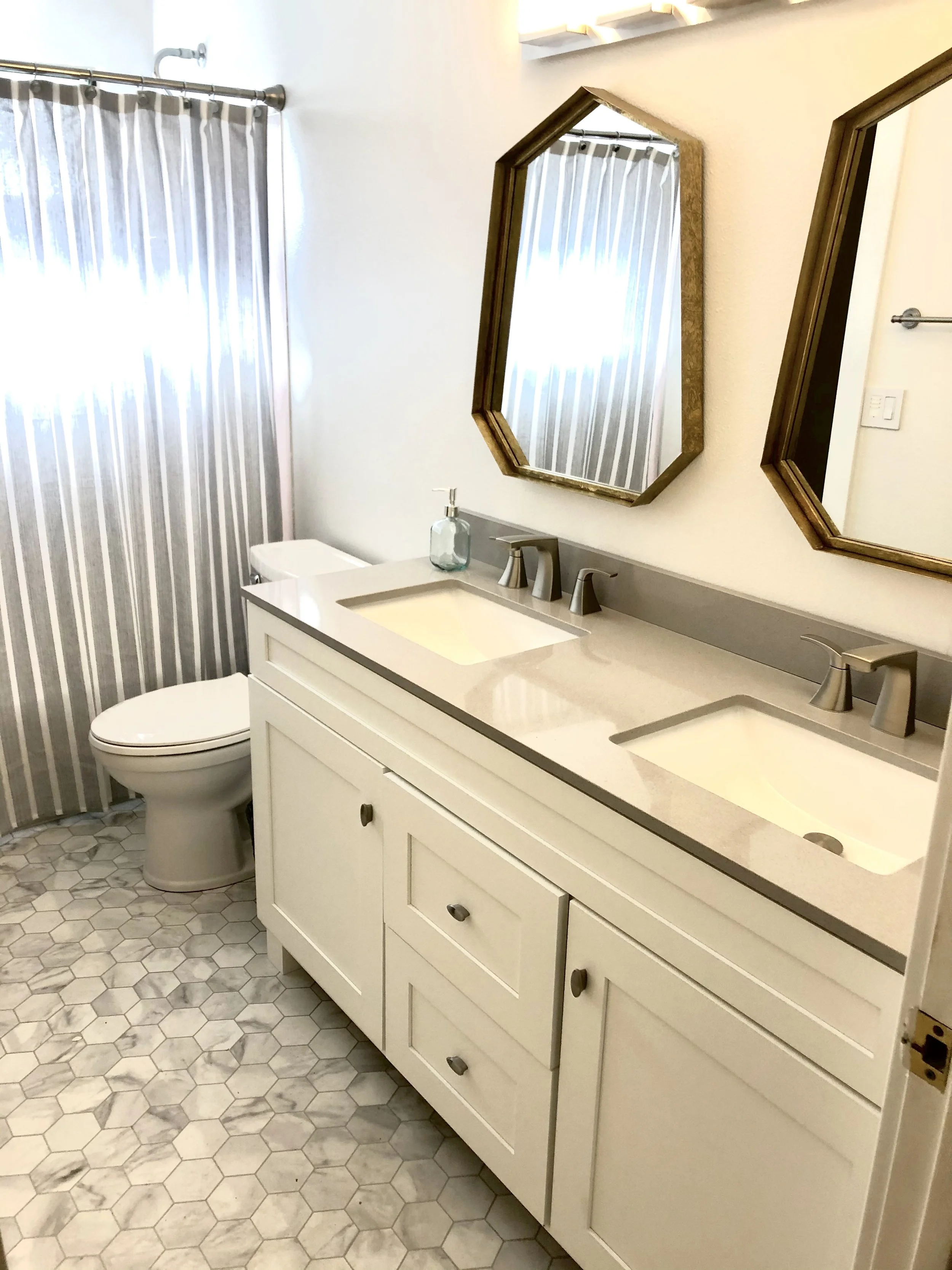 Modern bathroom with dual sinks and white cabinets, hexagonal tile flooring, striped shower curtain, and octagonal mirrors.