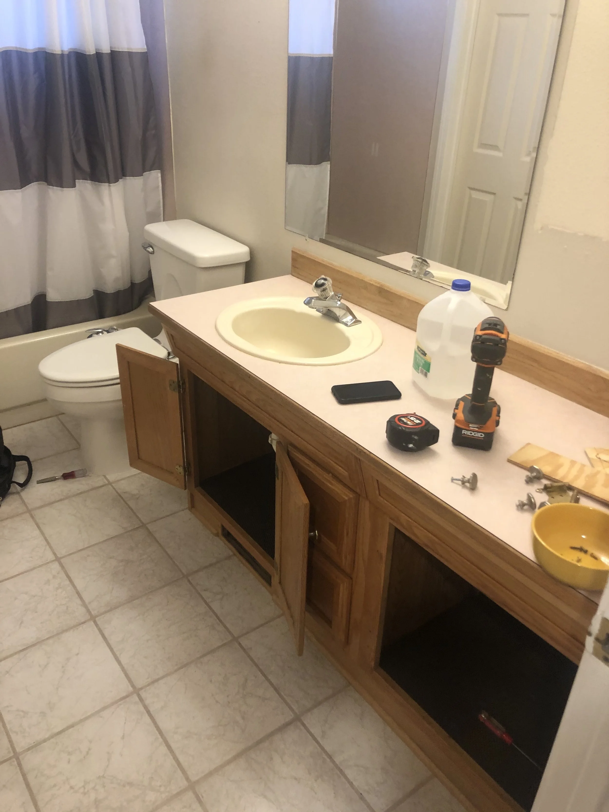 Bathroom with a sink, mirror, and wooden cabinet. The cabinet doors are open, exposing the interior. Various tools and items, including a drill, tape measure, screws, and a gallon jug, are scattered on the counter. A toilet and shower curtain are visible in the background.
