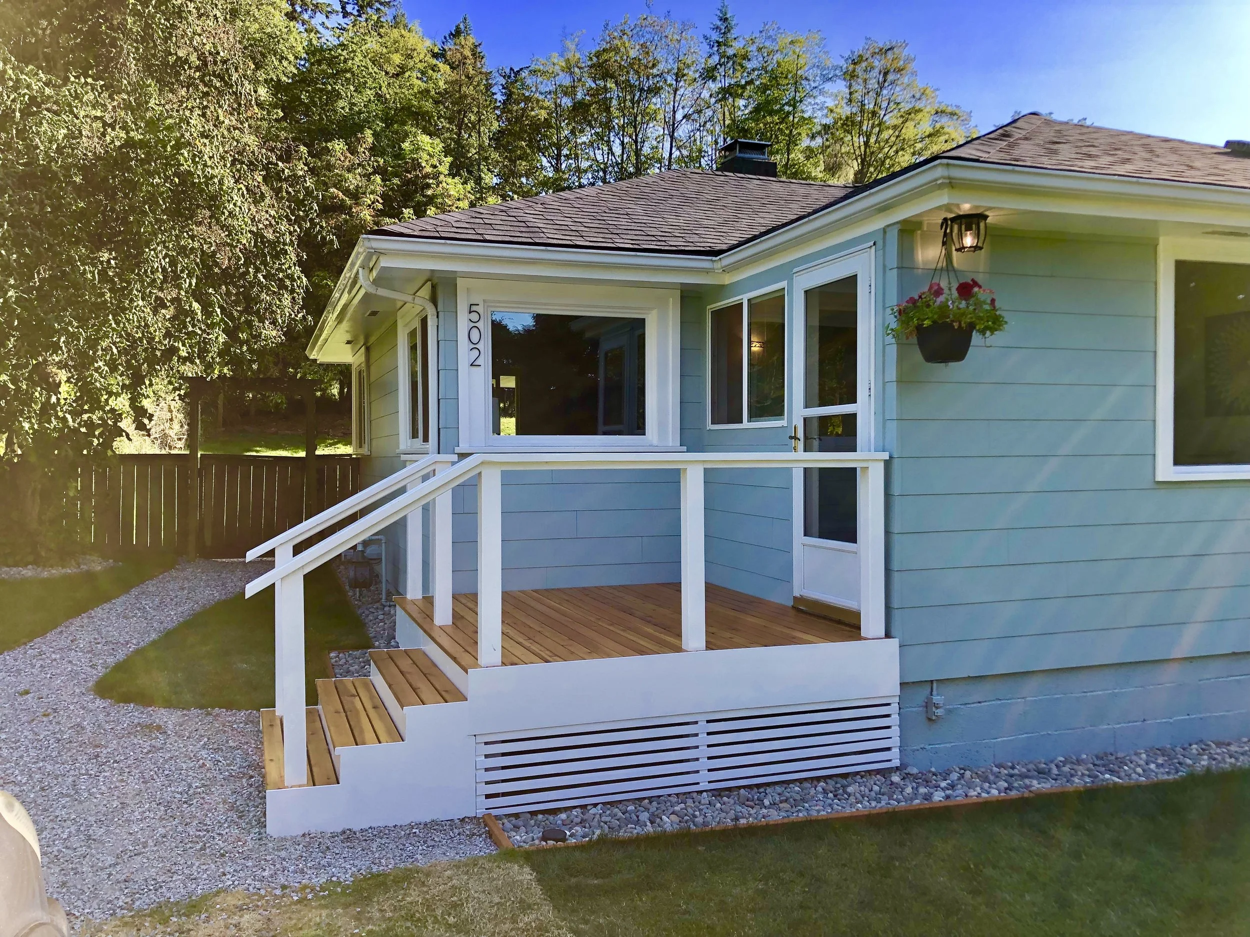 A light blue house with a small porch, white railings, and steps. The house number 502 is visible. There is a hanging flower pot near the door, and a gravel path leads to a wooden fence. The yard is grassy with trees in the background.