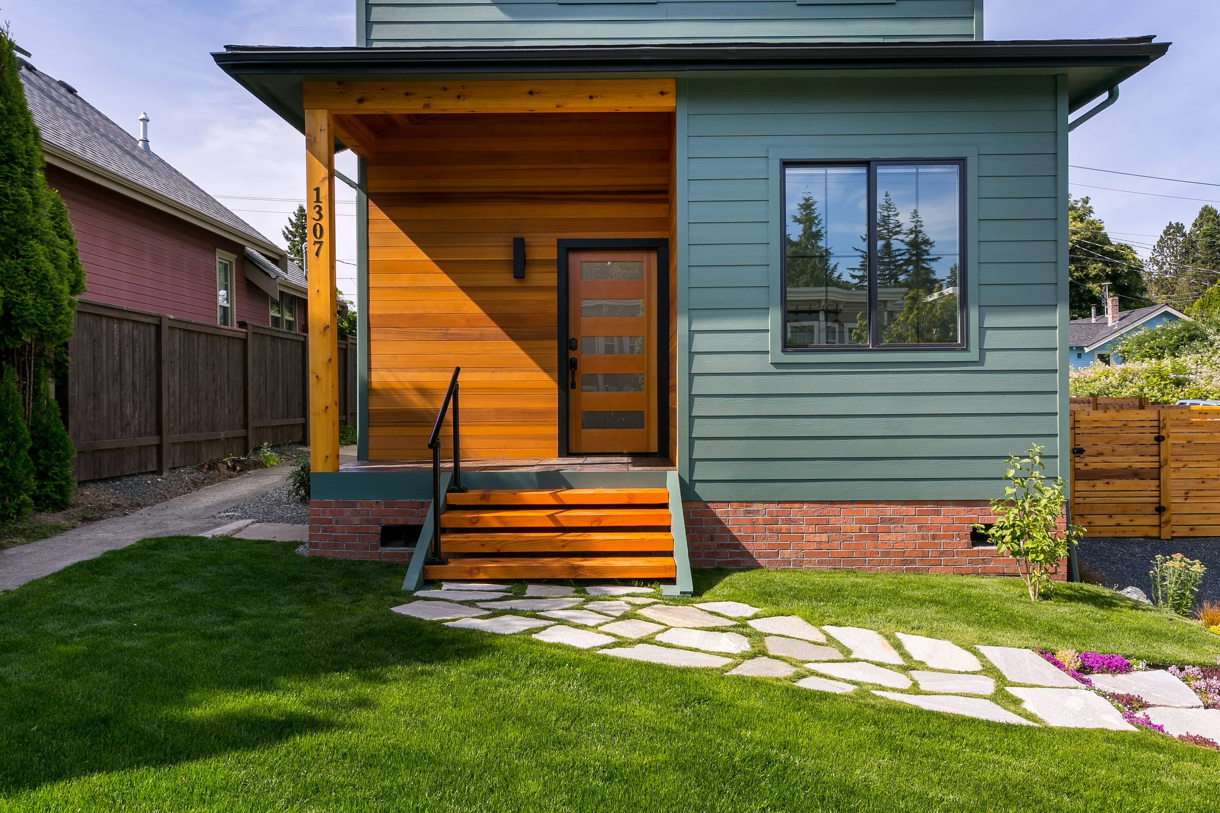 Front view of a modern residential house with a wooden and brick exterior, featuring a bright wood door, porch with stairs, a concrete walkway, and a green lawn in the foreground.