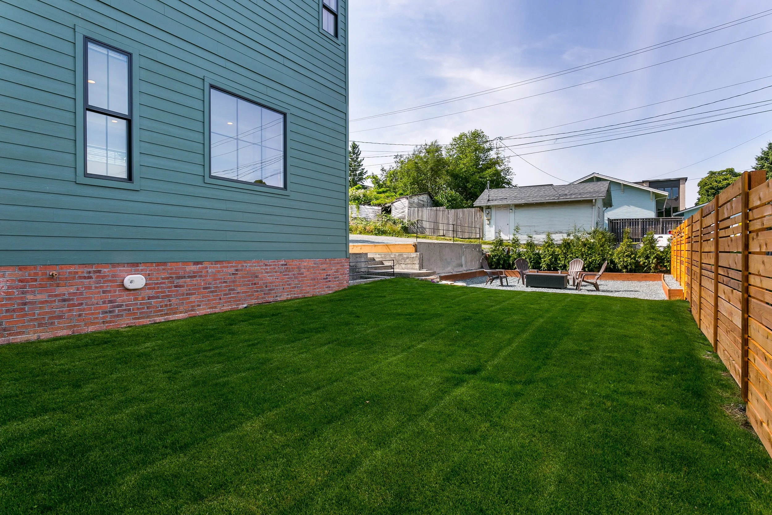 Backyard with green grass, a wooden fence, patio area with chairs, and a house with green siding and brick foundation
