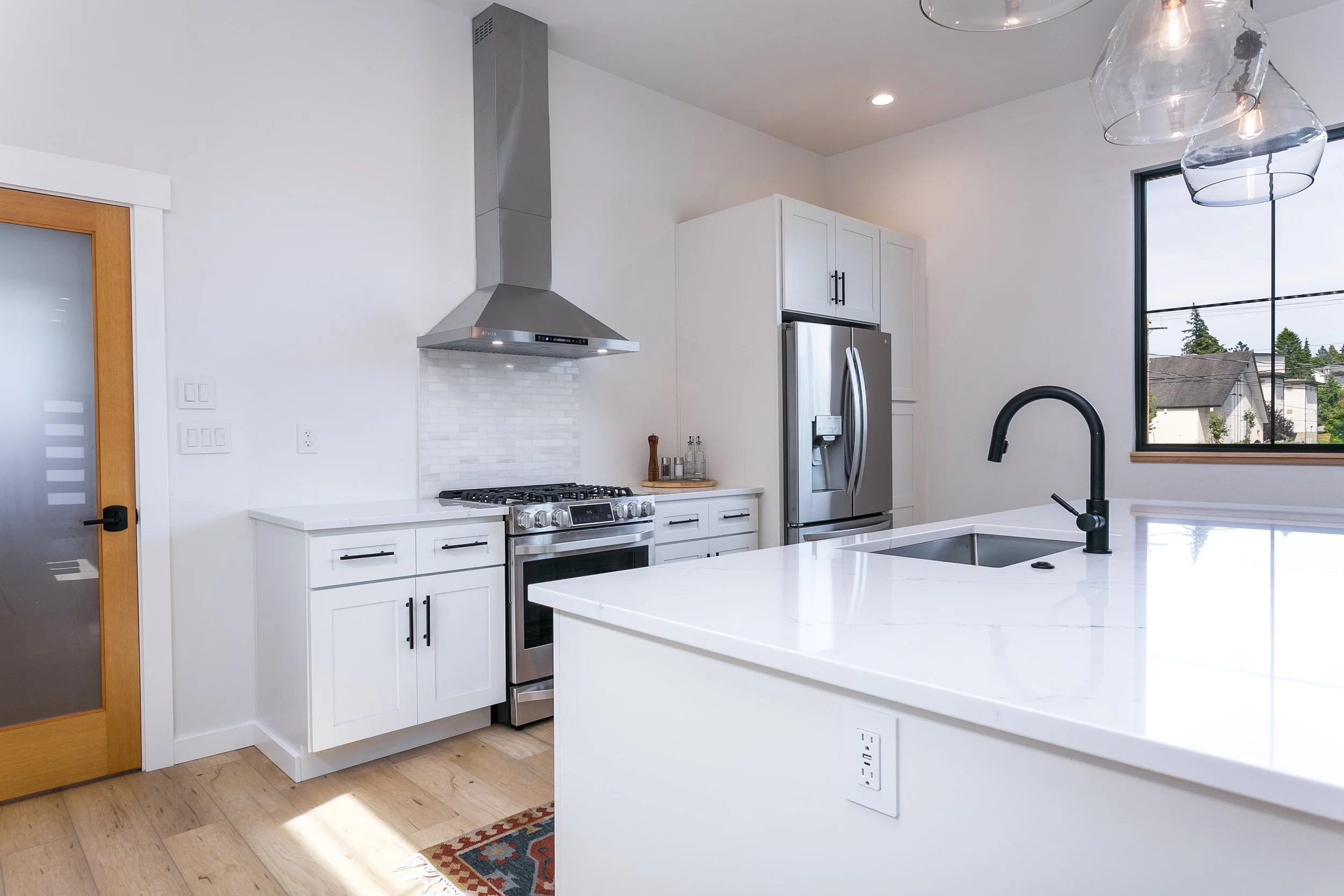 Modern kitchen with stainless steel appliances, white cabinets, black faucet, and large window.