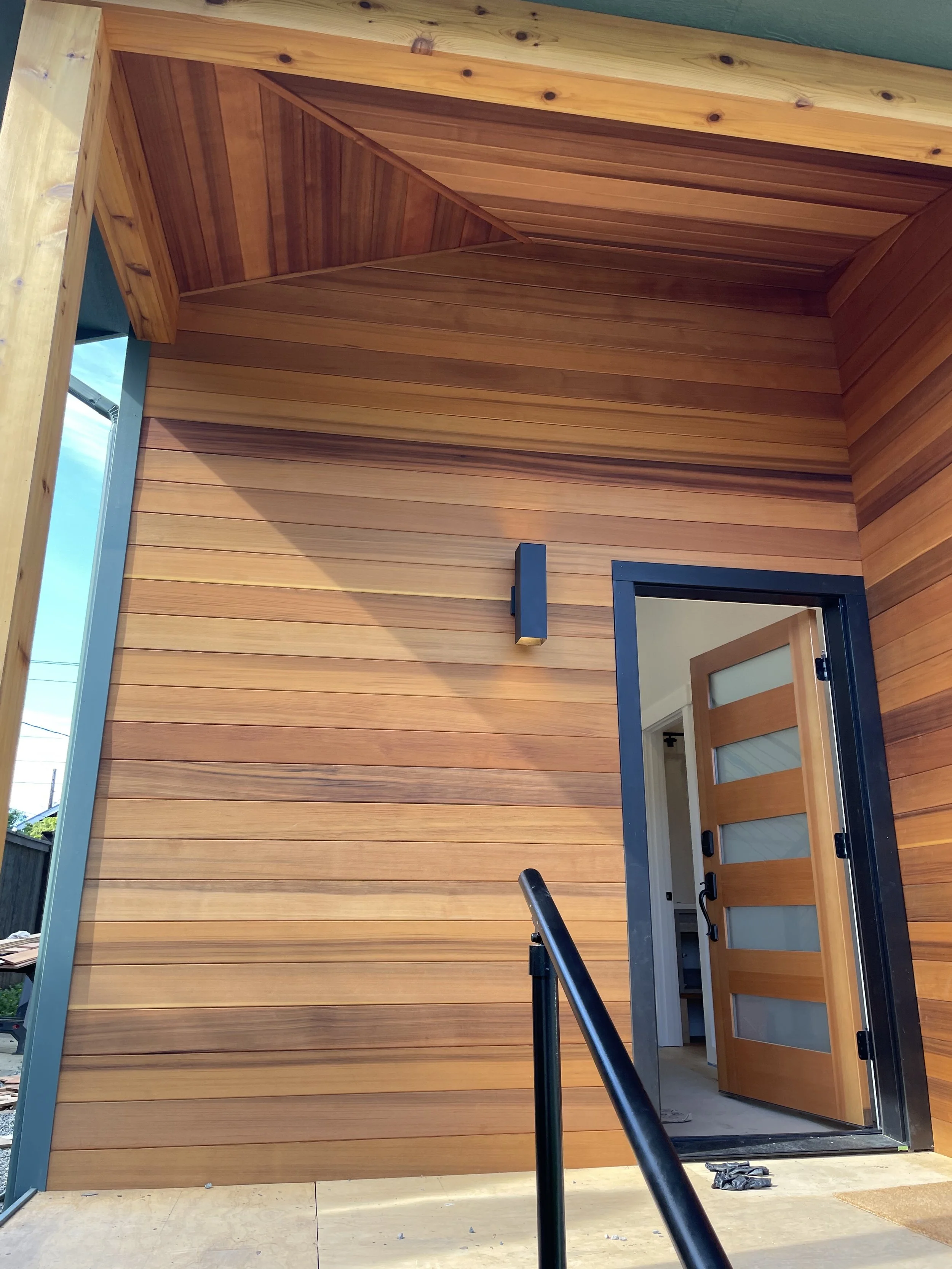 Wooden house exterior with a slatted wall and an open modern-style door with glass panels, black railing in the foreground.
