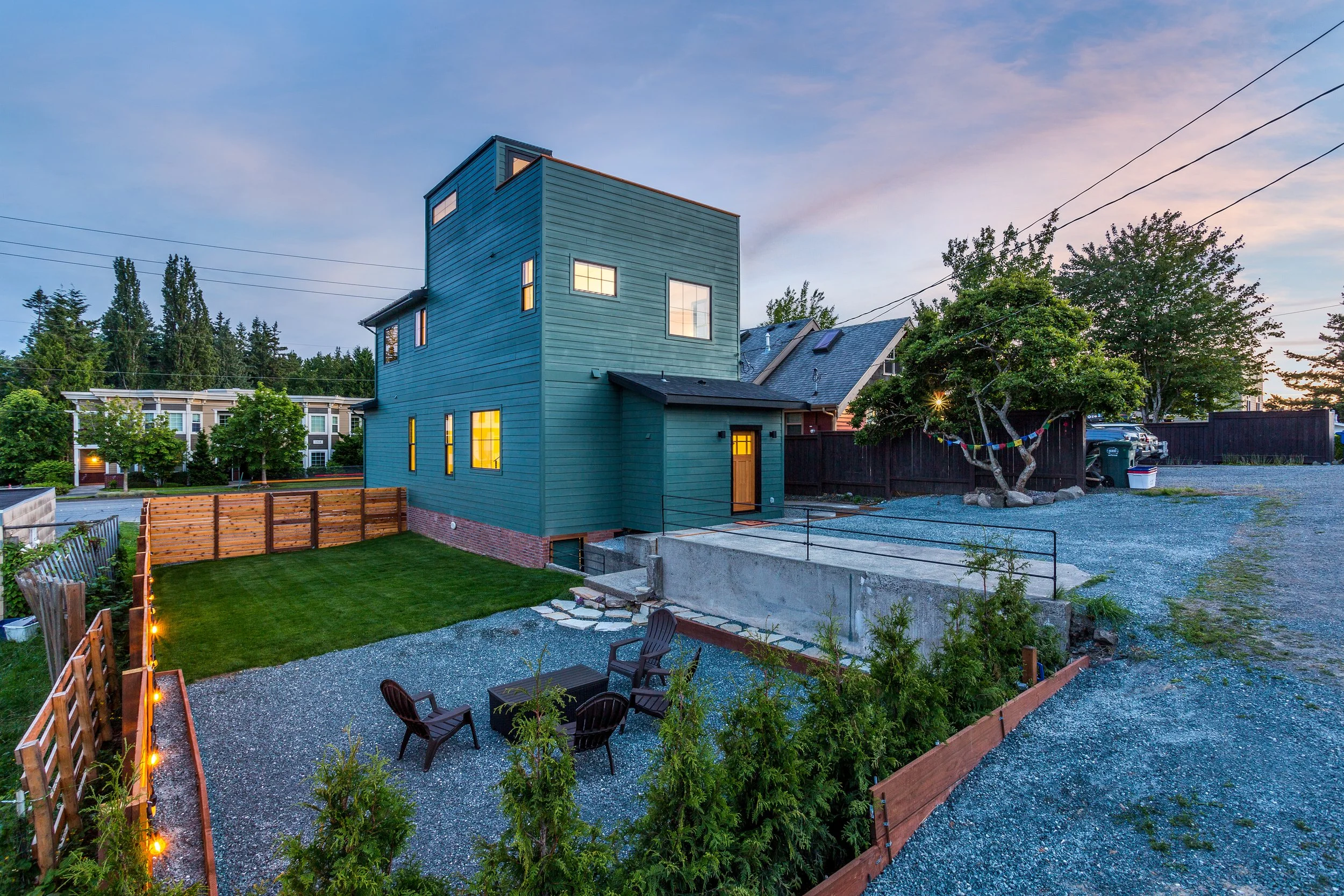 Modern two-story house with teal siding, large windows, and a sloped roof, set in a spacious yard with a gravel seating area, lawn, and wooden fence, surrounded by trees during sunset.