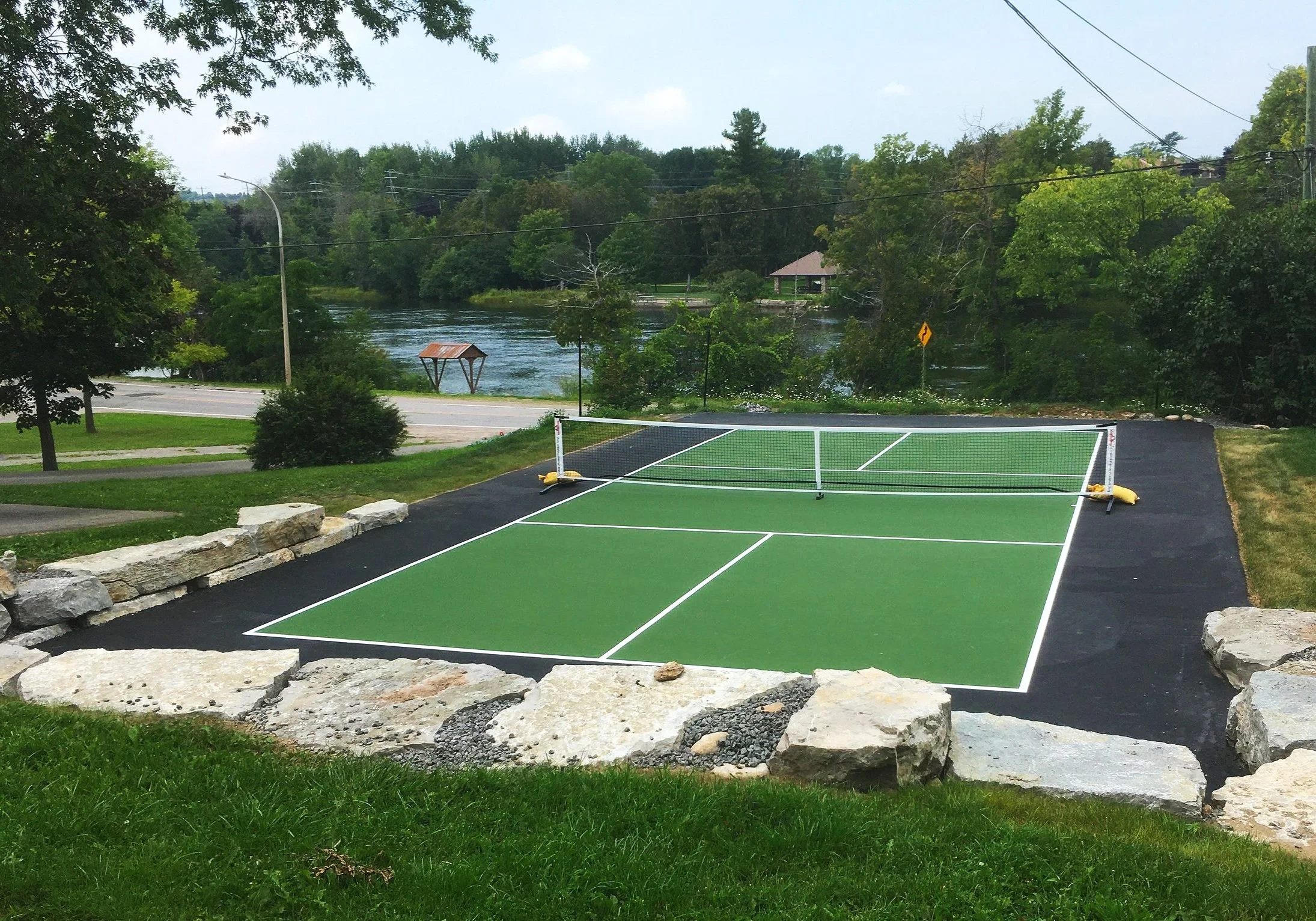 Outdoor pickleball court near a river, surrounded by trees and greenery.