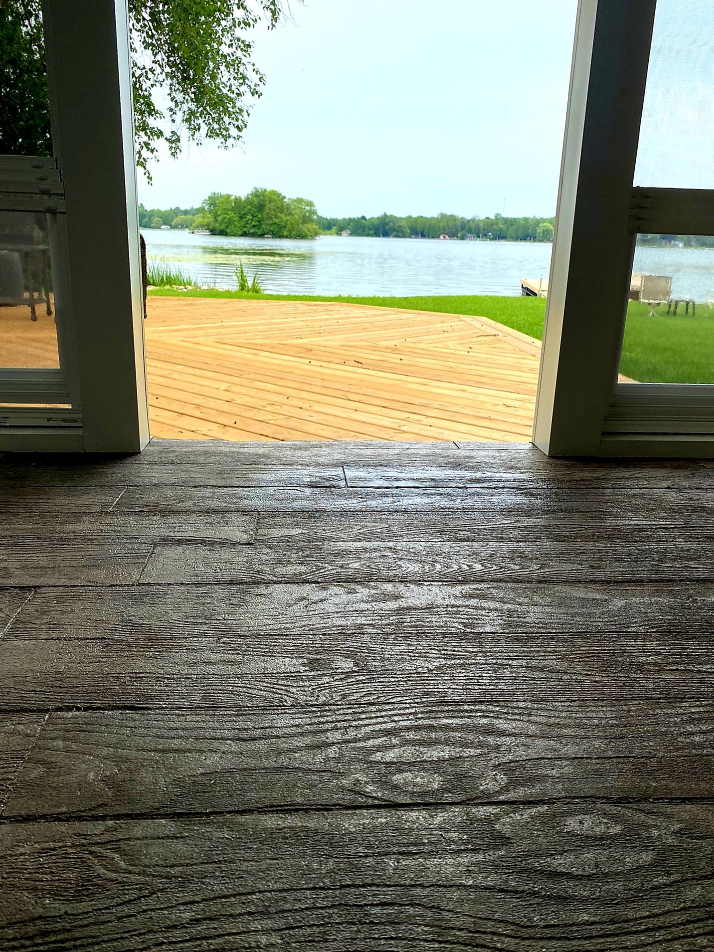 View through open doors showing a wooden deck, green grass, and a lake with trees in the background.