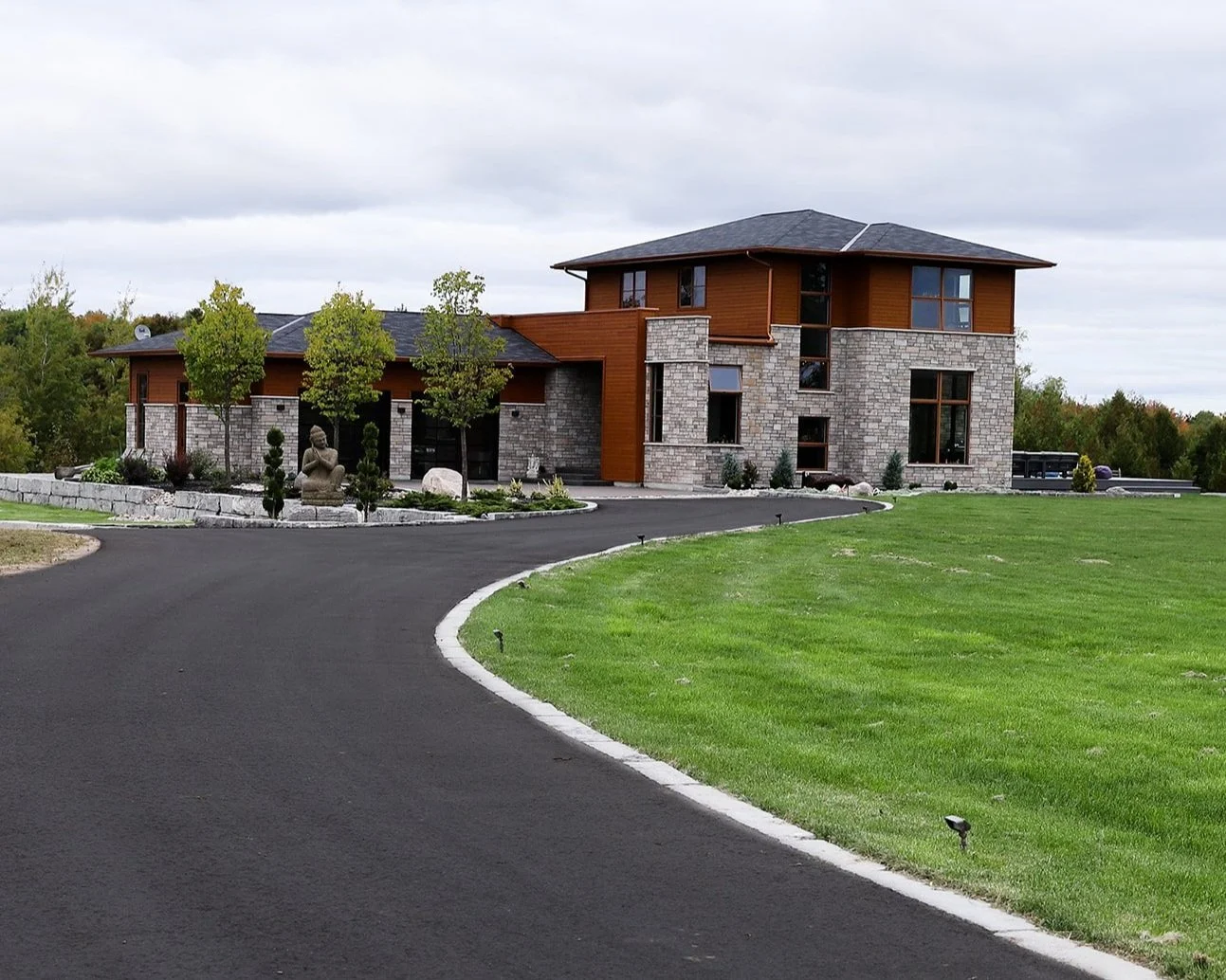 Modern house with stone and wood exterior, surrounded by green lawn, trees, and a curved asphalt paved driveway