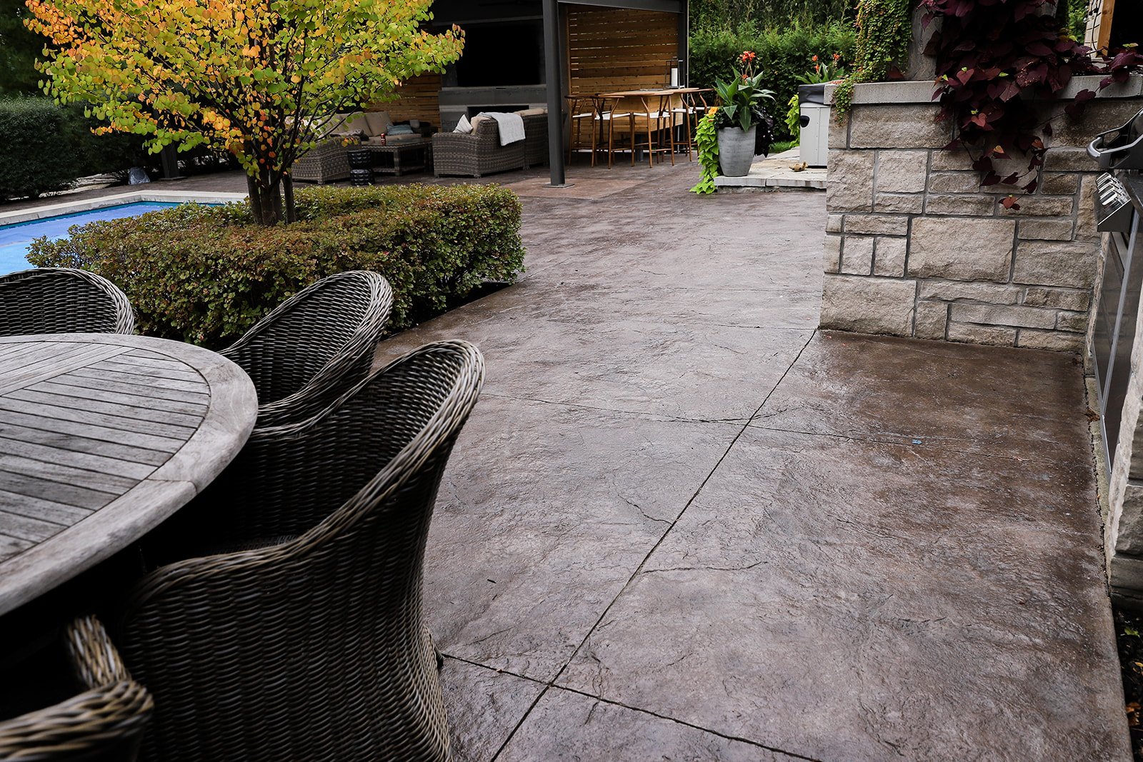 Backyard patio area with stone paving, outdoor furniture, round table with wicker chairs, small pool, greenery, and a covered seating area.