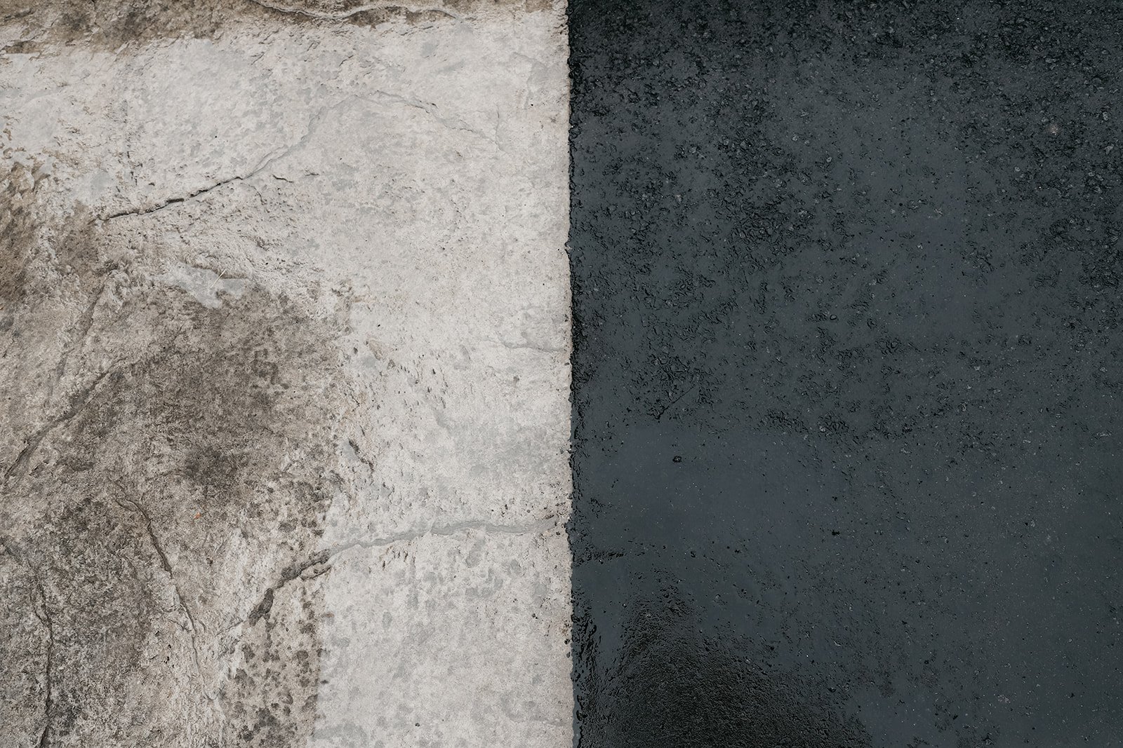 Close-up of a textured surface divided vertically, with light gray concrete on the left and darker, wet asphalt on the right.