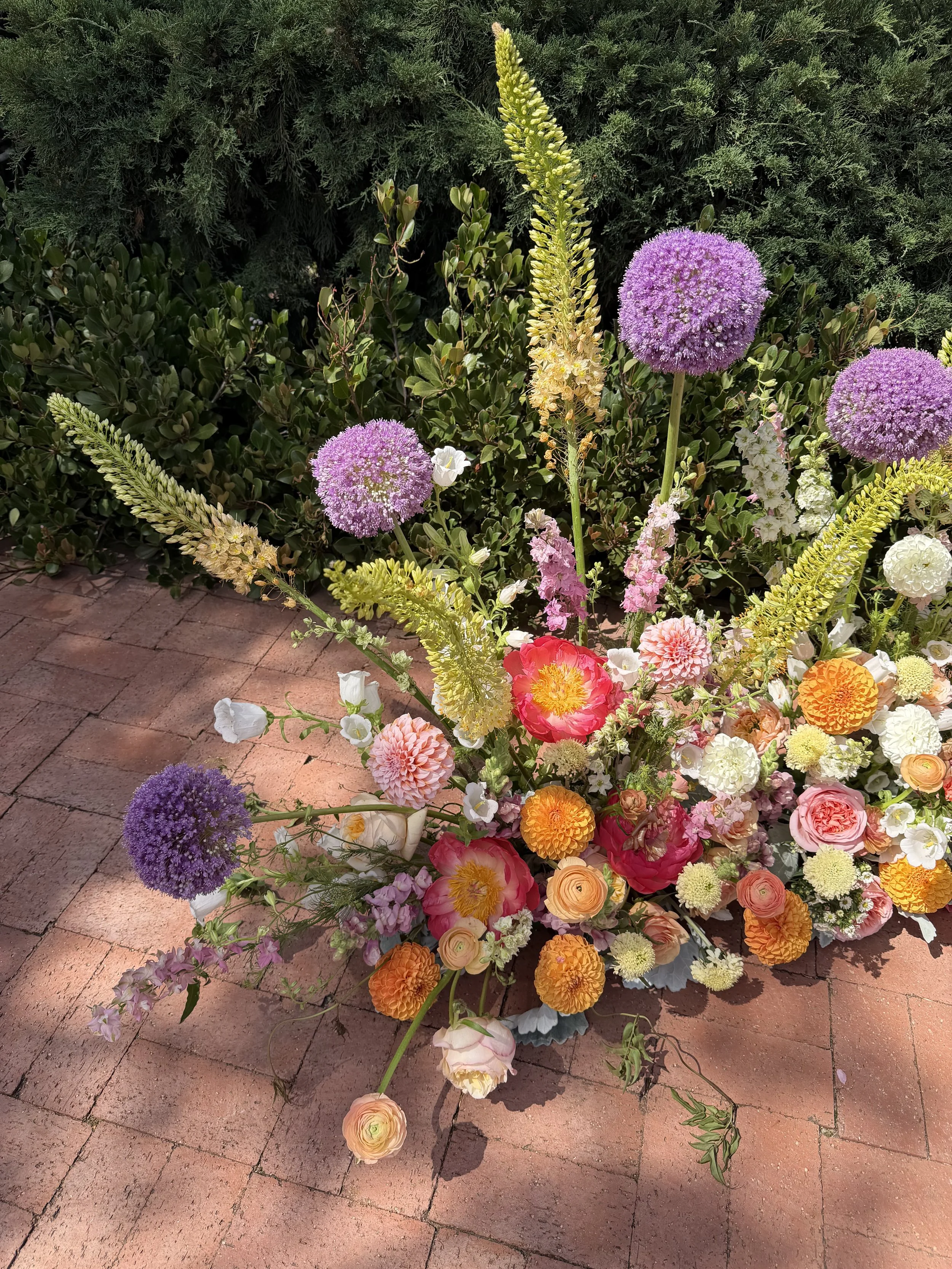 An arrangement of colorful flowers on a brick pathway, including purple alliums, pink roses, orange marigolds, white flowers, and other assorted blooms, with green bushes and trees in the background.