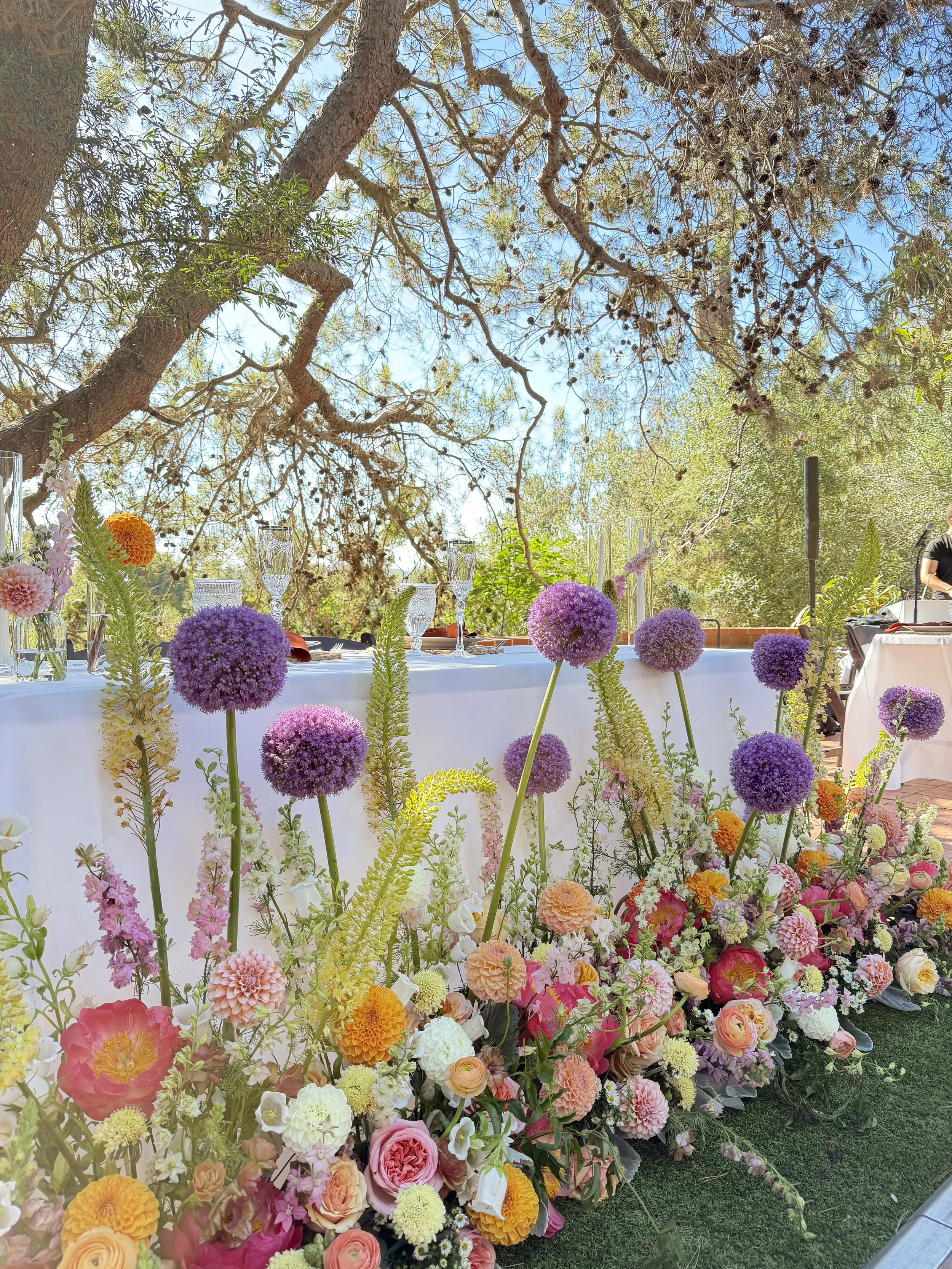 Colorful flower arrangement on a table outdoors under trees and blue sky, with glasses and tableware visible, likely at a celebration or event.