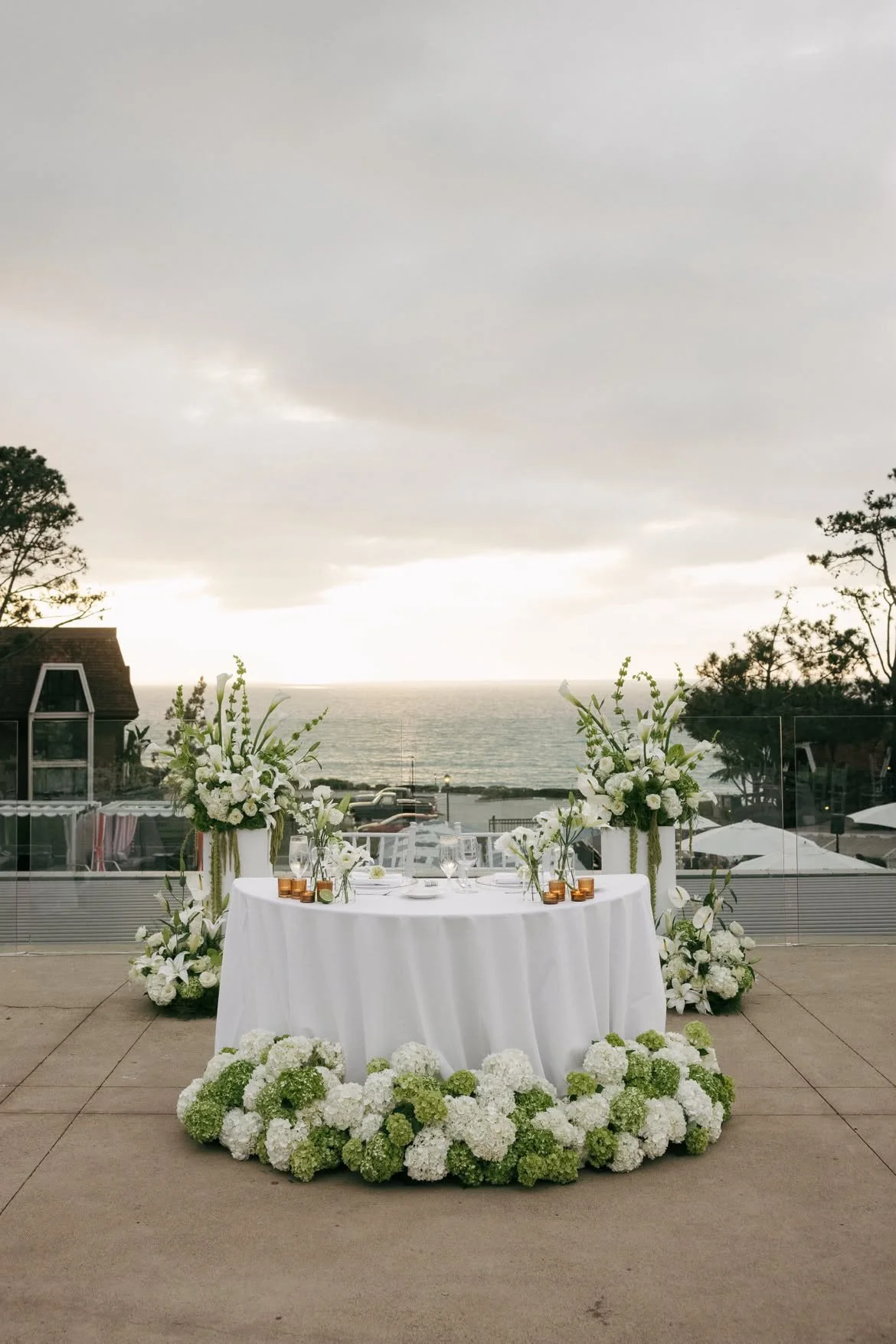 Wedding reception table decorated with white flowers and candles, set outdoors with a view of the ocean and a cloudy sky in the background.